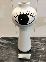 Front view of white ceramic vase with large black eye design facing forward, showing the shape and pedestal base