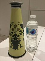 View of the tall conical yellow pottery vase with black and brown hand-painted floral designs beside a water bottle for scale.