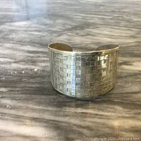 Front view of a silver-tone metal cuff bracelet with a basket weave pattern on a grey marble surface.
