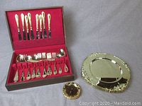 Overall view of the open flatware chest with gold-plated flatware arranged inside, along with one charger plate and one small plate/coaster placed beside the box.