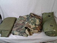 Photo showing all folded items: solid green wool blankets, two folded poncho liners in camouflage pattern, and rolled green tent in its pouch.
