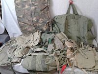Military bags and harnesses displayed on a flat surface, featuring various canvas messenger bags, travel bags, harnesses, and felt fabric, mostly in olive green and camouflage patterns.