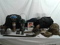 Photo shows assortment of military and camping gear on white cloth background. Items include canteens, lanterns, sleeping bags, helmets, caps, belts, and a cologne bottle.