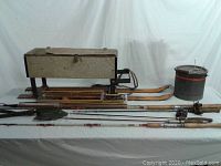 Full lot view showing ice fishing tackle box on skis, fishing rods laid in front, collapsible shovel, and metal bucket.