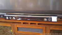 Samsung soundbar positioned under a TV on a wooden stand, showing front view with speaker drivers visible.