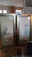 Wide shot showing both framed prints side by side. Each has a gold frame with black inner trim. The image shows the full length of the prints with nature and floral imagery.