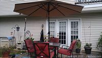Outdoor patio set showing glass and metal square table with four matching metal-frame chairs with red cushions, taupe patio umbrella fully extended over table, placed on a patio next to house.