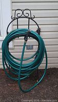 Wrought iron hose stand holding a green garden hose with attached nozzle, positioned outside against a house wall.