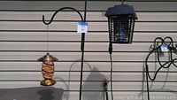 Photo of the full wrought iron shepherd's hook with hanging glass hummingbird feeder on left hook and electric bug zapper on right hook against beige siding background.