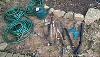 Photo showing three green garden hoses coiled on the ground alongside a green plastic watering can, Kobalt hatchet, wand hose attachment, hedge clippers, long-handled pruners, auger bit, and sprinkler.