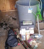 View of grey plastic tote bin with landing net leaning, small tackle box on top, assorted fishing rods and lures on ground