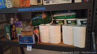 Shelf holding packages of toilet paper, paper towels, plastic cutlery, lunch bags, and other paper products.