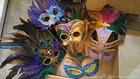 Five Marie-Gras style masks with feathers and sequins in purple, teal, blue, gold, and pink colors