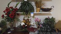 Overview showing silk red roses in clear glass vase, various mixed silk plants and floral arrangements including orchids, and decorative containers.