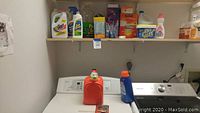 Laundry detergent red and blue jugs on washing machine with shelves above containing cleaning supplies