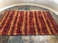 Photo of a 5x7 shaggy area rug placed on tiled floor with a striped pattern of red, orange, and yellow colors.