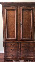 Front view of the solid wood armoire showing closed paneled doors and six drawers below with ornate handles.