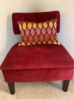 Small red upholstered chair with wooden legs and a decorative pillow with multicolored leaf pattern showing front and upper part of chair.
