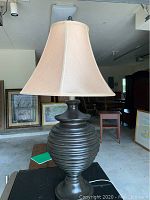 Photo of a table lamp with a brown ribbed wooden base and beige square fabric shade on a black surface in a room with framed artwork in the background
