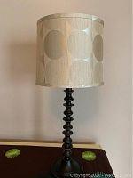 Photo of a table lamp featuring a black wooden spindle base and a beige fabric shade with a large circle pattern.