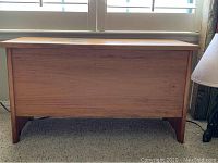 Small natural wood chest photographed from front showing rectangular shape, flat top, and four solid legs on carpeted floor near window.