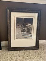 Framed sepia-toned print featuring a wooden beach fence with shadows on sandy beach, inside rustic dark wood frame with glass cover.