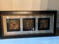 Large rectangular frame in black with gold trim containing three square medallion pieces with floral designs in gold tone on black background.