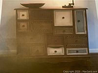 Frontal view showing asymmetrical arrangement of 13 drawers in brown and off-white with bronze knob pulls and scalloped base