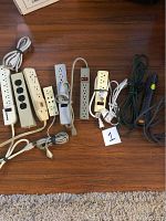 Top down view of 10 extension cords and power strips arranged on a wood floor, showing various colors and types including cords with switches and multiple outlets.