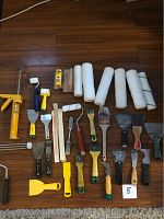 Overview of painting tools including caulking gun, various paint rollers with covers, several paint brushes, scrapers, putty knives, painter's tape, and mixing stick on wood floor.