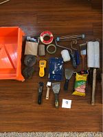 Full layout of all painting supplies on wood floor showing orange painting tray, rollers, brushes, scrapers, steel wool, tapes, masking paper, moisture meter, and spackling compound.
