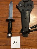 Front view of diving knife with serrated blade and textured black handle, displayed next to the black plastic sheath case with belt straps.