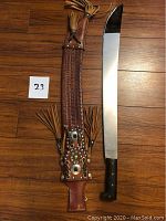 Full view of machete with leather sheath showing blade length and sheath decorations with tassels