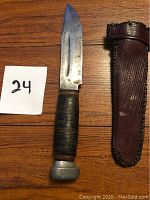 Top view of antique knife with leather-wrapped handle and metal pommel, placed next to brown textured leather sheath on wood surface