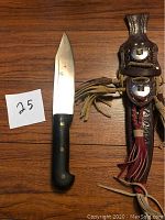 Machete blade and black handle with sheath side by side on wooden surface, labelled '25'.