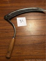 Photo showing the full sickle blade with curved blade and attached wooden handle on wooden surface.