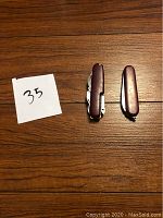 Two Swiss Army knives side by side on wooden surface, with a paper note marked '35'. One closed, one partially opened showing several tools.