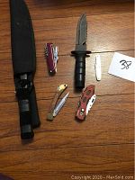 Six knives arranged on wood floor: a large fixed blade hunting knife with black sheath, a red Swiss Army-style knife, a small white folding utility knife, a small brass-handled folding knife, and two other folding knives with patterned handles.
