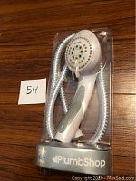 Full view of the new PlumbShop shower head and its hose inside clear plastic packaging on a wooden surface.