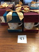 Eight Godiva gift mugs in single packaging boxes, each tied with gold Godiva ribbon, displayed on a wood surface.