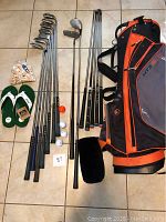 Wide shot showing all golf equipment including clubs, shafts, golf bag, golf balls, tees, and golf sandals arranged on floor tiles.