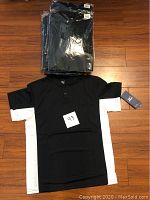 Nine men's Mizuno baseball shirts, mostly in sealed plastic packaging, laid on a wood floor. One shirt removed from packaging shown flat with black body and white side panels, short button placket collar, tags attached.