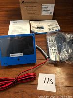 American Dynamics Public View Monitor kit including the monitor with blue housing, remote control in plastic bag, power cable, box, and user manuals on wooden surface.