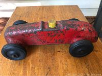 Pressed steel toy race car resting on wood surface, showing red painted body with wear and brass-colored knob on top.