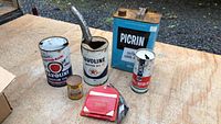 Top-down photo showing seven vintage automotive oil and petrol cans including a large Havoline can, Picrin can, and smaller cans on a wooden surface outdoors.