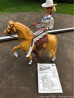 Barbie doll riding a yellow/tan horse with bridle and saddle. Paperwork is visible in front.