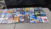 Top view of 26 DVD movies laid out on table. Variety of family, comedy, and adventure titles visible, some sealed in original packaging.