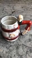 Ceramic mug shaped like a barrel with red painted bands, featuring a sculpted man forming the handle, seen from the side.