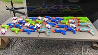 A collection of 37 assorted Nerf guns displayed on a table outdoors. The guns vary in style and color including blue, green, white, and gray with orange tips.