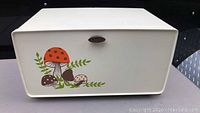 Front angled view of the cream colored metal bread box showing mushroom graphics and wooden handle.